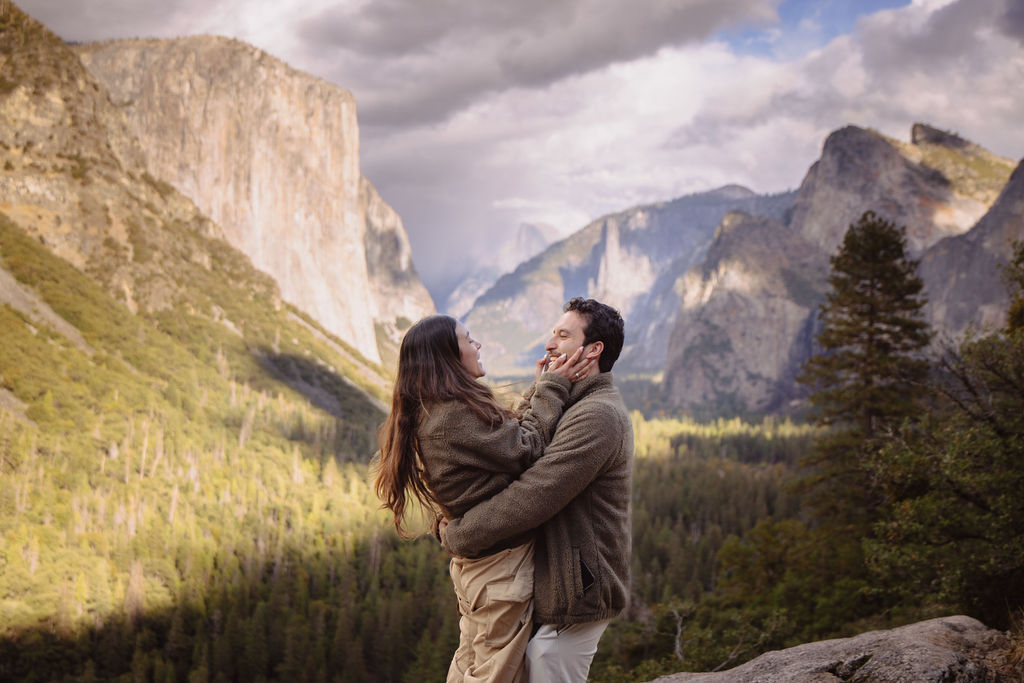 A couple smiles at each other while embracing, with a scenic view of mountains and a forest in the background under a cloudy sky taken by a yosemite proposal photographer