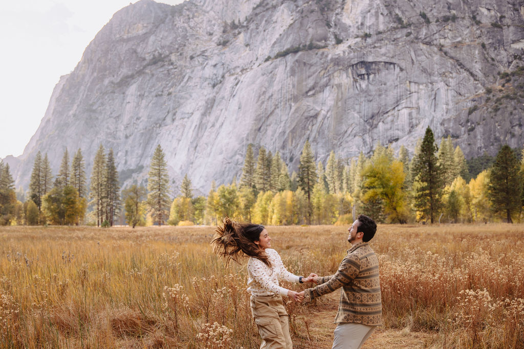 A couple stands in a sunlit field surrounded by trees, looking at each other affectionately. Sunlight creates a warm, golden glow taken by a yosemite proposal photographer