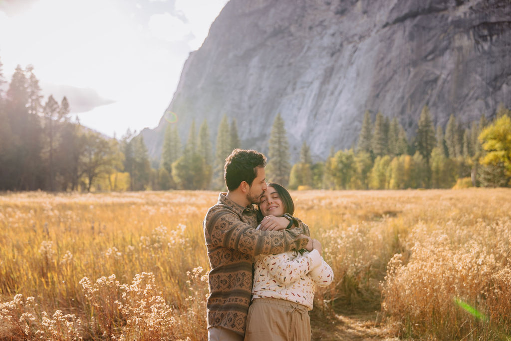 A couple stands in a sunlit field surrounded by trees, looking at each other affectionately. Sunlight creates a warm, golden glow taken by a yosemite proposal photographer