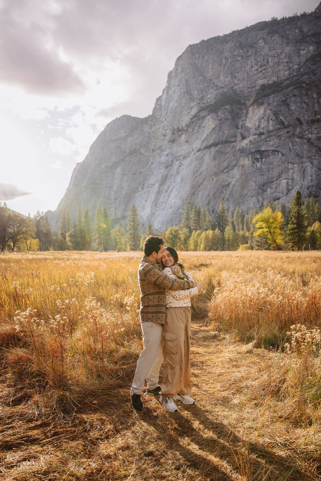 A couple stands in a sunlit field surrounded by trees, looking at each other affectionately. Sunlight creates a warm, golden glow taken by a yosemite proposal photographer