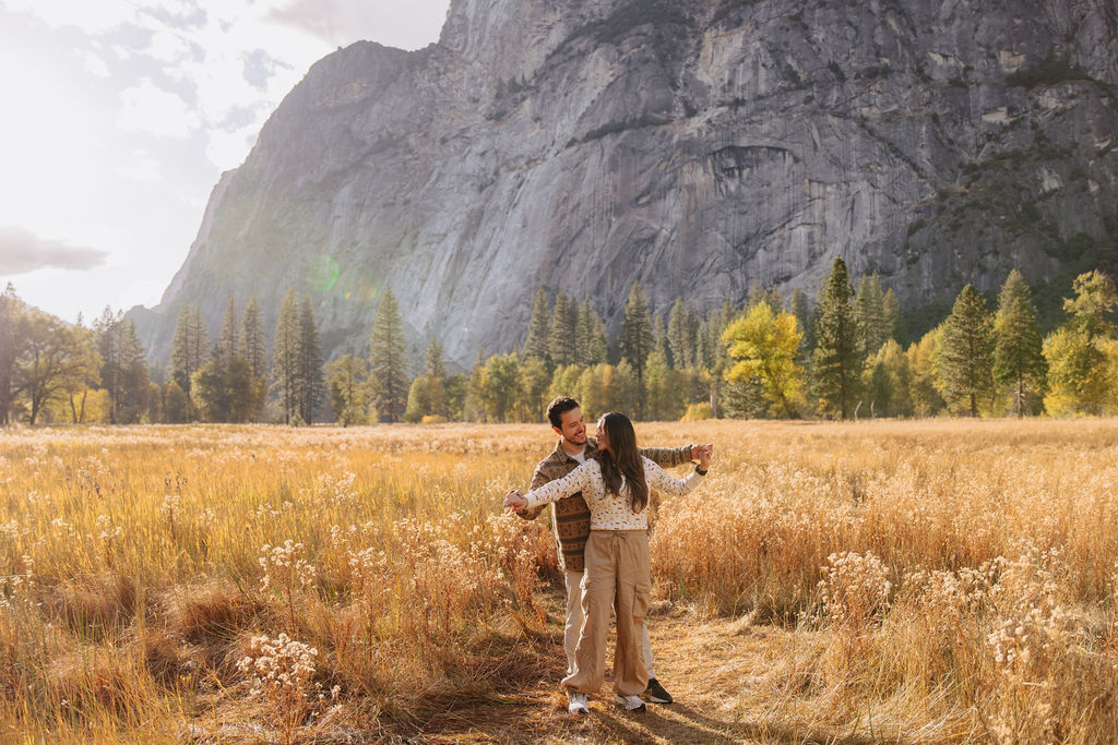 A couple stands in a sunlit field surrounded by trees, looking at each other affectionately. Sunlight creates a warm, golden glow taken by a yosemite proposal photographer