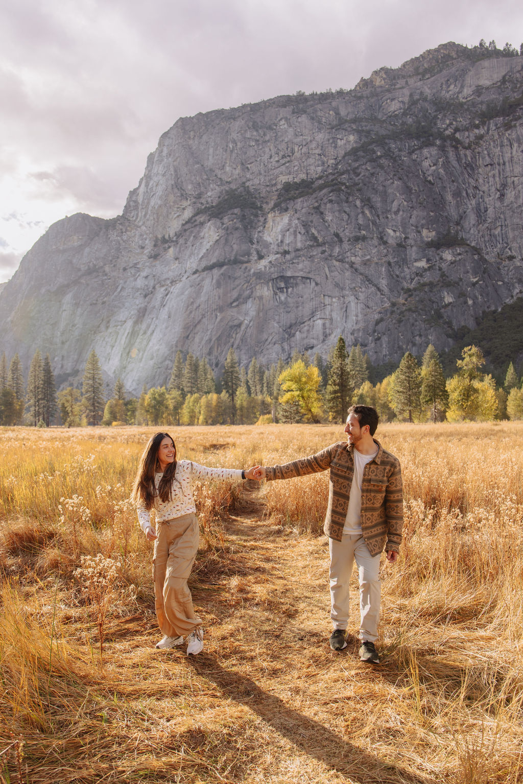 A couple stands in a sunlit field surrounded by trees, looking at each other affectionately. Sunlight creates a warm, golden glow taken by a yosemite proposal photographer