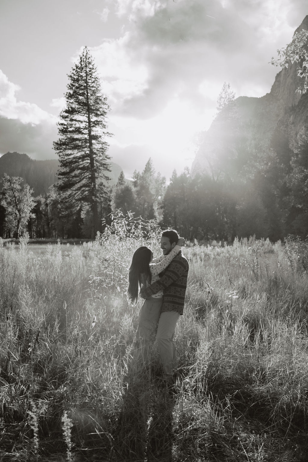 A couple stands in a sunlit field surrounded by trees, looking at each other affectionately. Sunlight creates a warm, golden glow taken by a yosemite proposal photographer