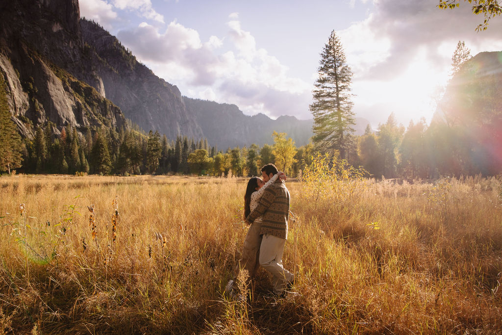 A couple stands in a sunlit field surrounded by trees, looking at each other affectionately. Sunlight creates a warm, golden glow taken by a yosemite proposal photographer