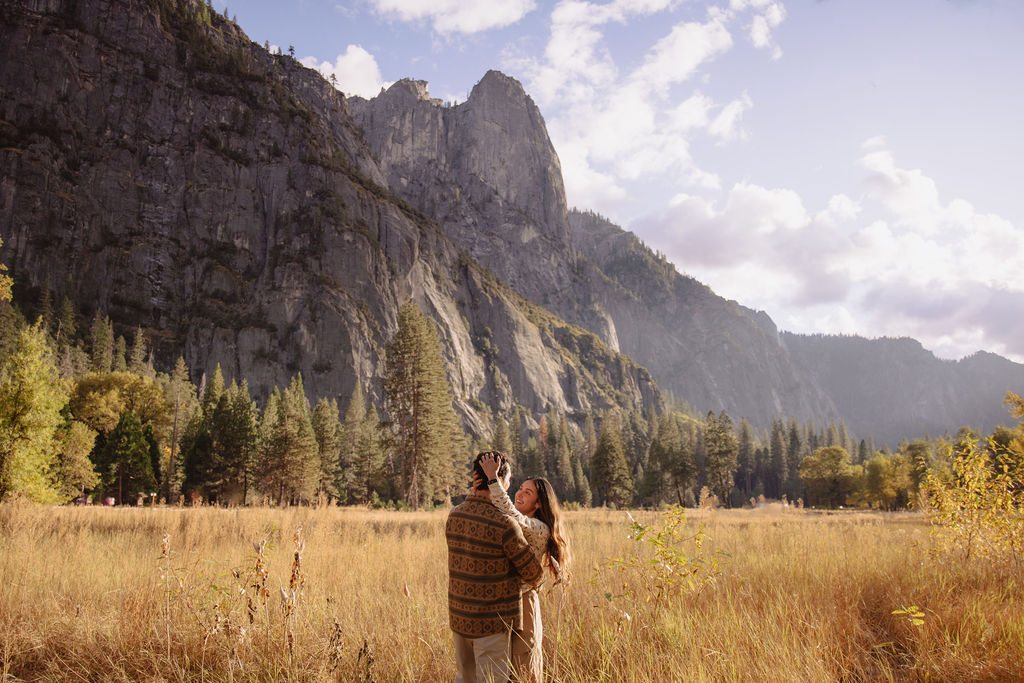 A couple stands in a sunlit field surrounded by trees, looking at each other affectionately. Sunlight creates a warm, golden glow taken by a yosemite proposal photographer