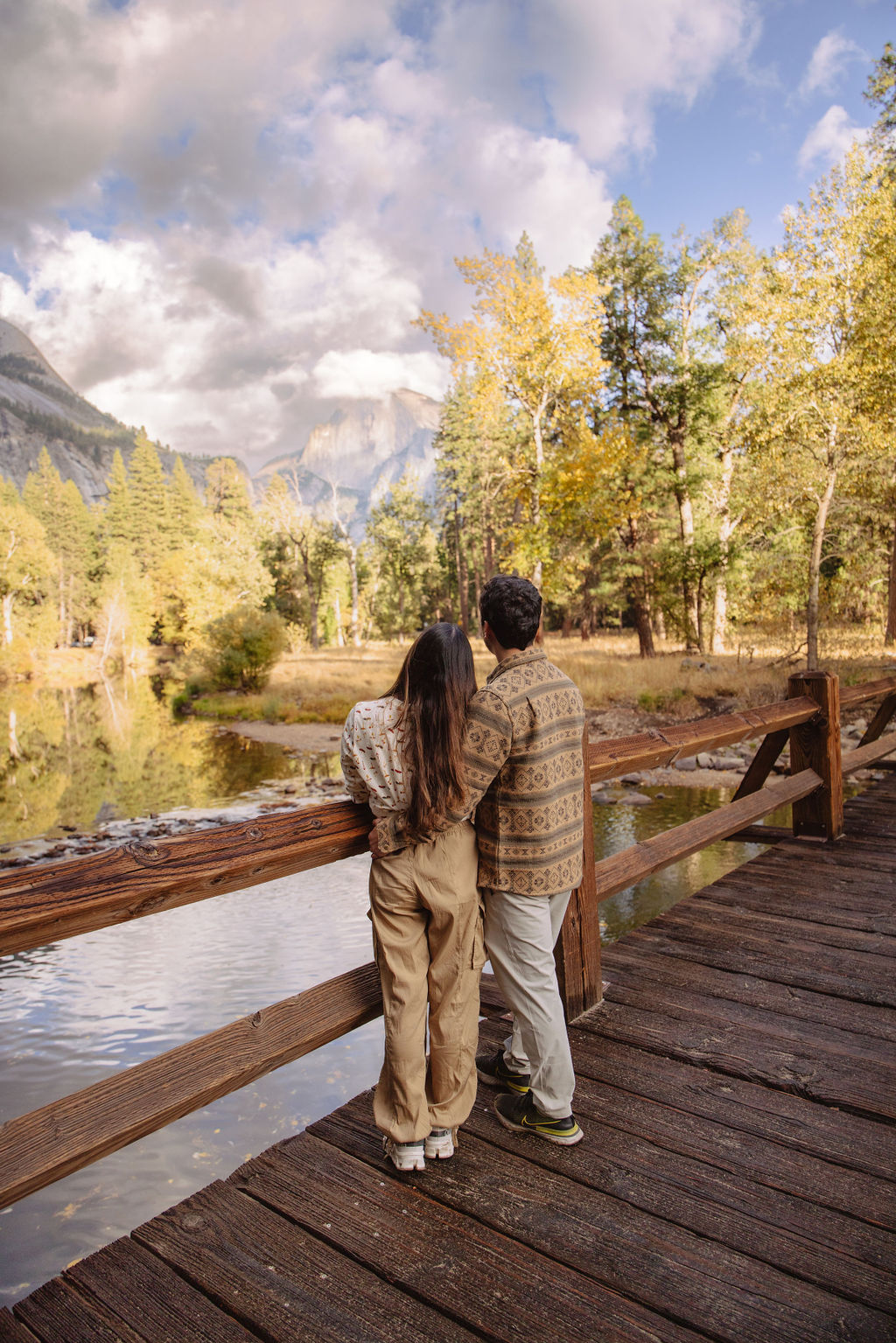 A couple stands in a sunlit field surrounded by trees, looking at each other affectionately. Sunlight creates a warm, golden glow taken by a yosemite proposal photographer