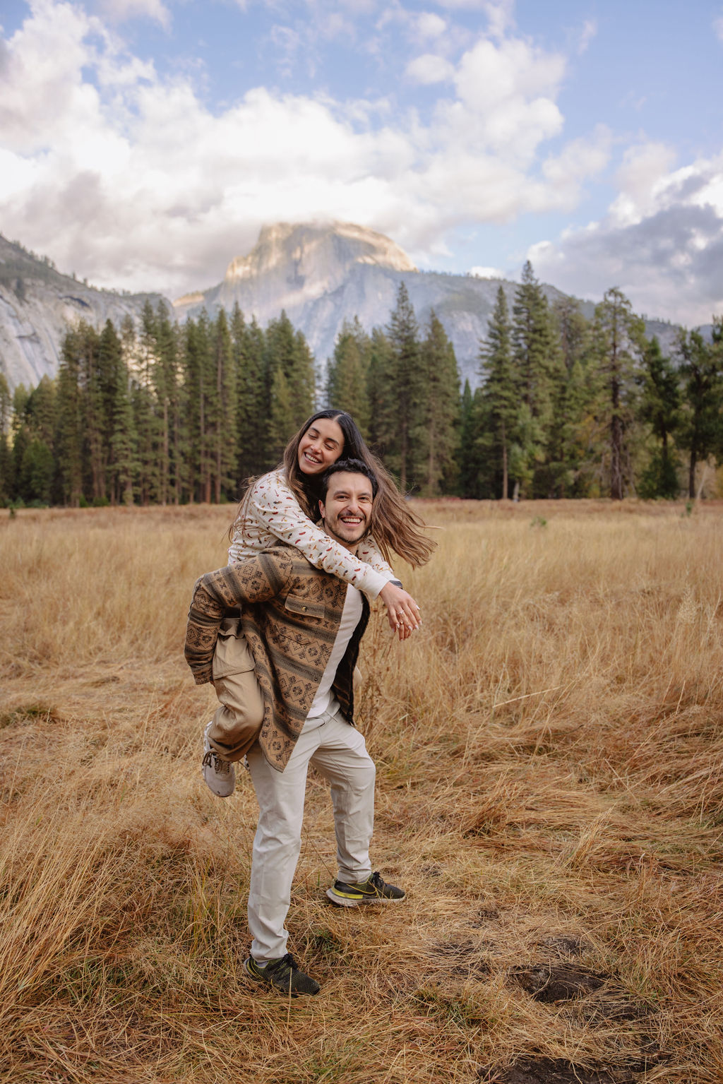 A couple stands in a sunlit field surrounded by trees, looking at each other affectionately. Sunlight creates a warm, golden glow taken by a yosemite proposal photographer