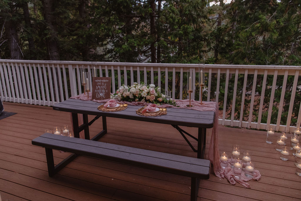 A picnic table on a wooden deck is decorated with flowers, candles, and a pink tablecloth, surrounded by trees and additional candles on the ground.