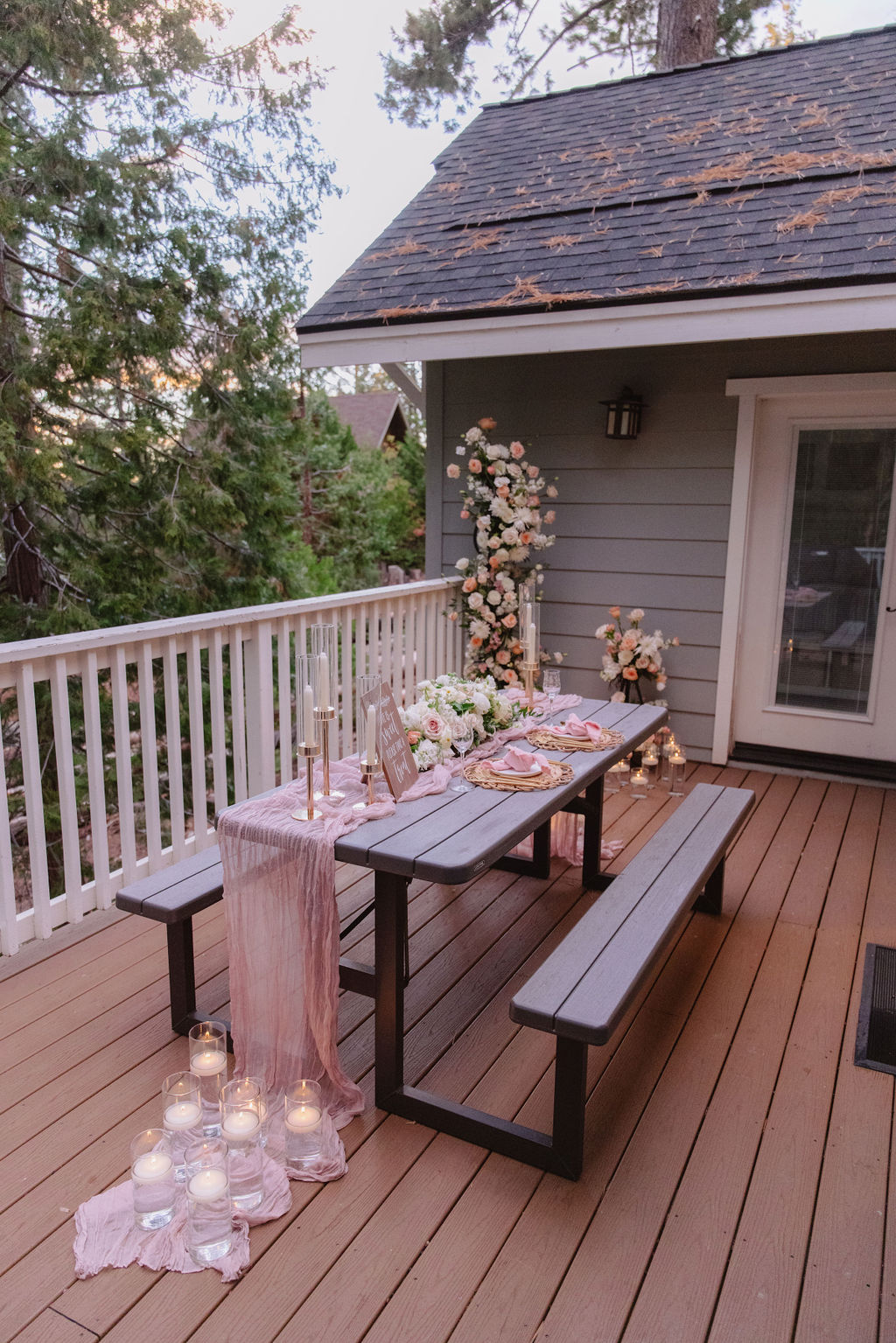 A picnic table on a wooden deck is decorated with flowers, candles, and a pink tablecloth, surrounded by trees and additional candles on the ground.