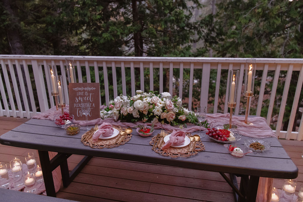 A picnic table on a wooden deck is decorated with flowers, candles, and a pink tablecloth, surrounded by trees and additional candles on the ground.