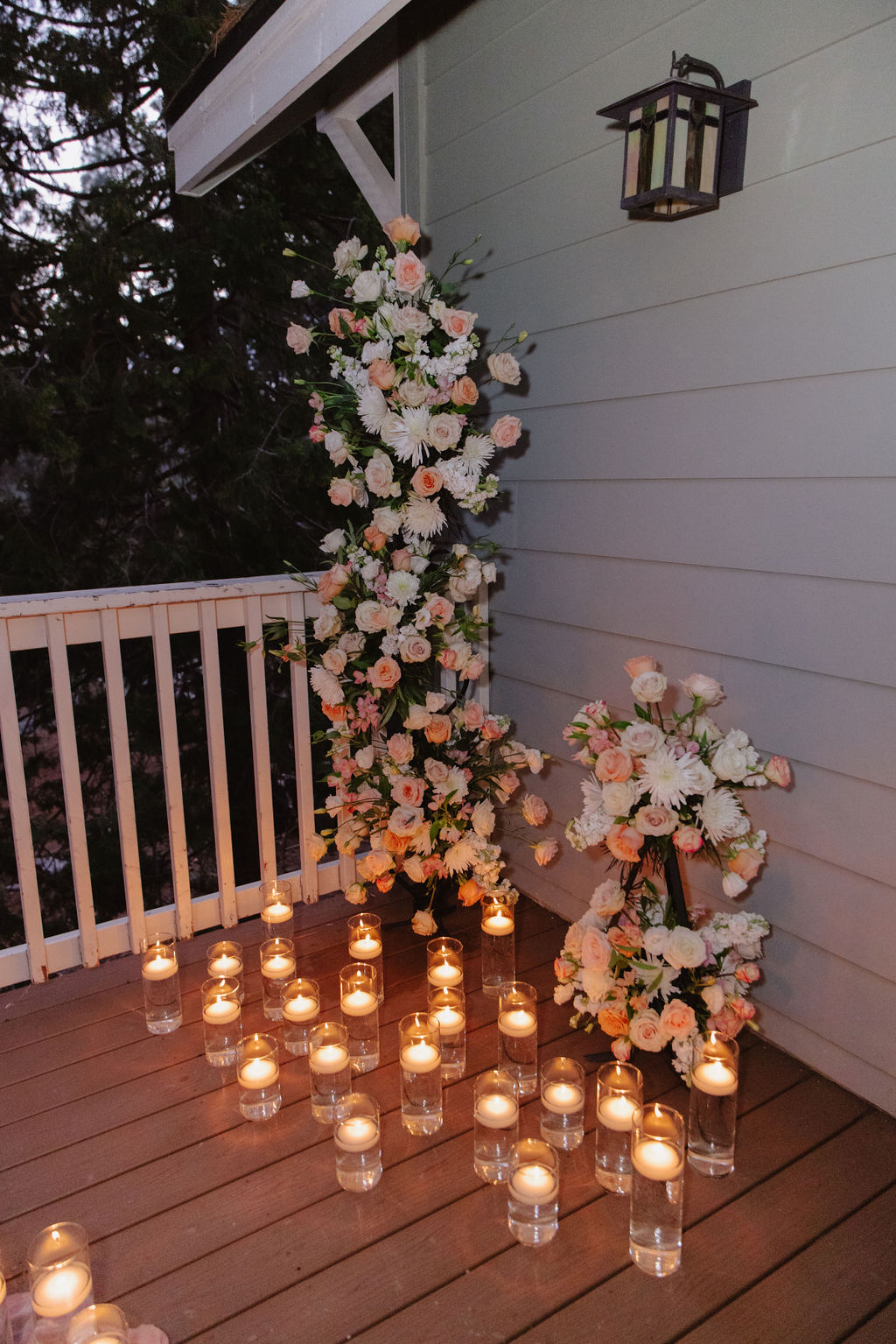 A picnic table on a wooden deck is decorated with flowers, candles, and a pink tablecloth, surrounded by trees and additional candles on the ground.