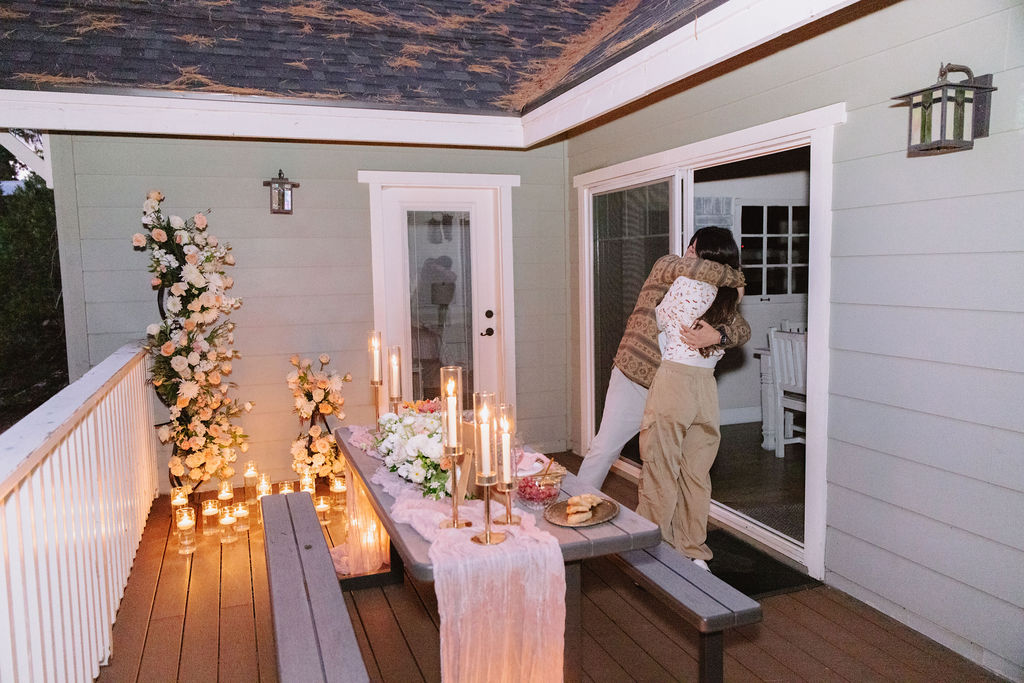 A couple embraces on a patio decorated with candles, flowers, and a table set for a meal.