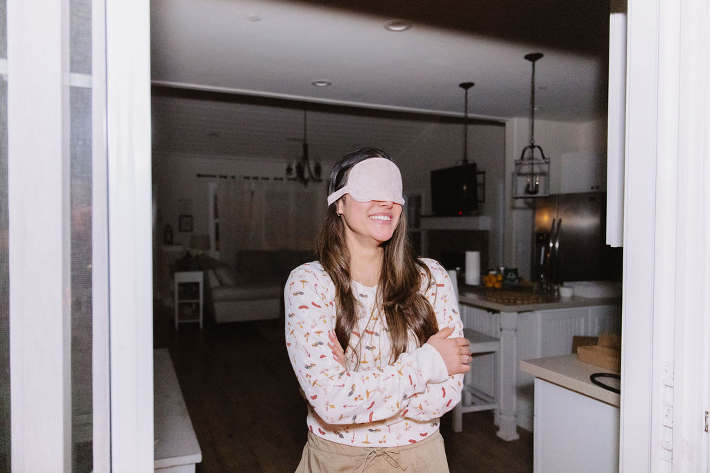 A man smiles while holding a stack of photos behind a woman wearing a blindfold. They are indoors.