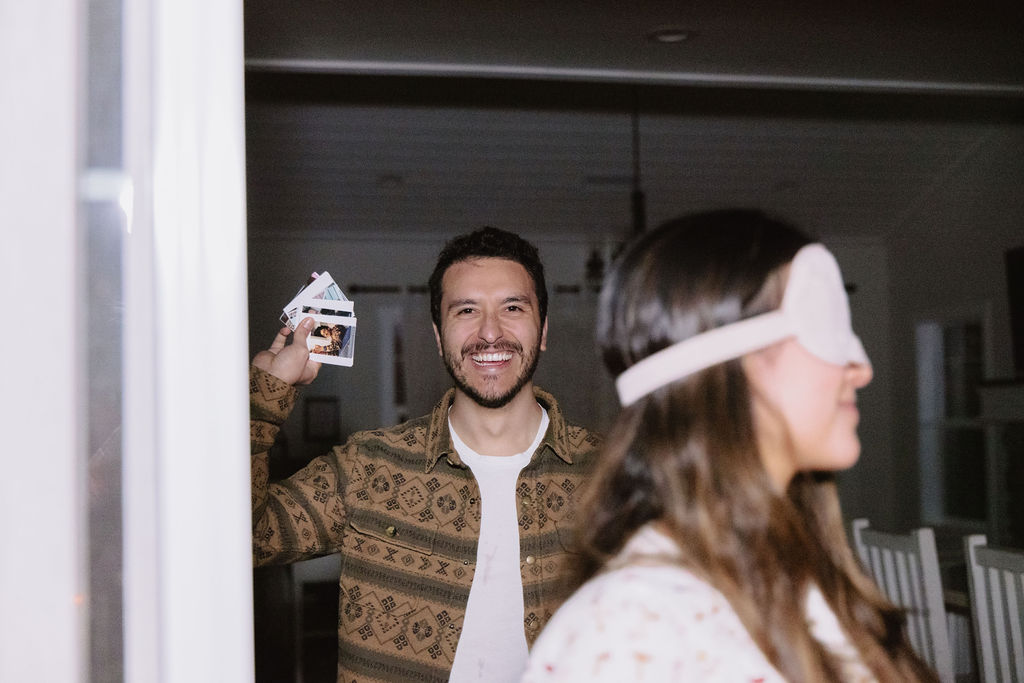 A man smiles while holding a stack of photos behind a woman wearing a blindfold. They are indoors.