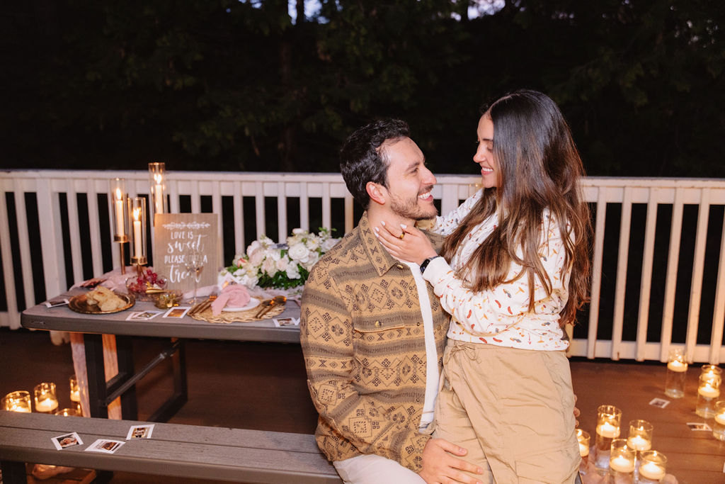 A couple sits on a picnic table, surrounded by candles and flowers, looking at each other. A sign and snacks are in the background.