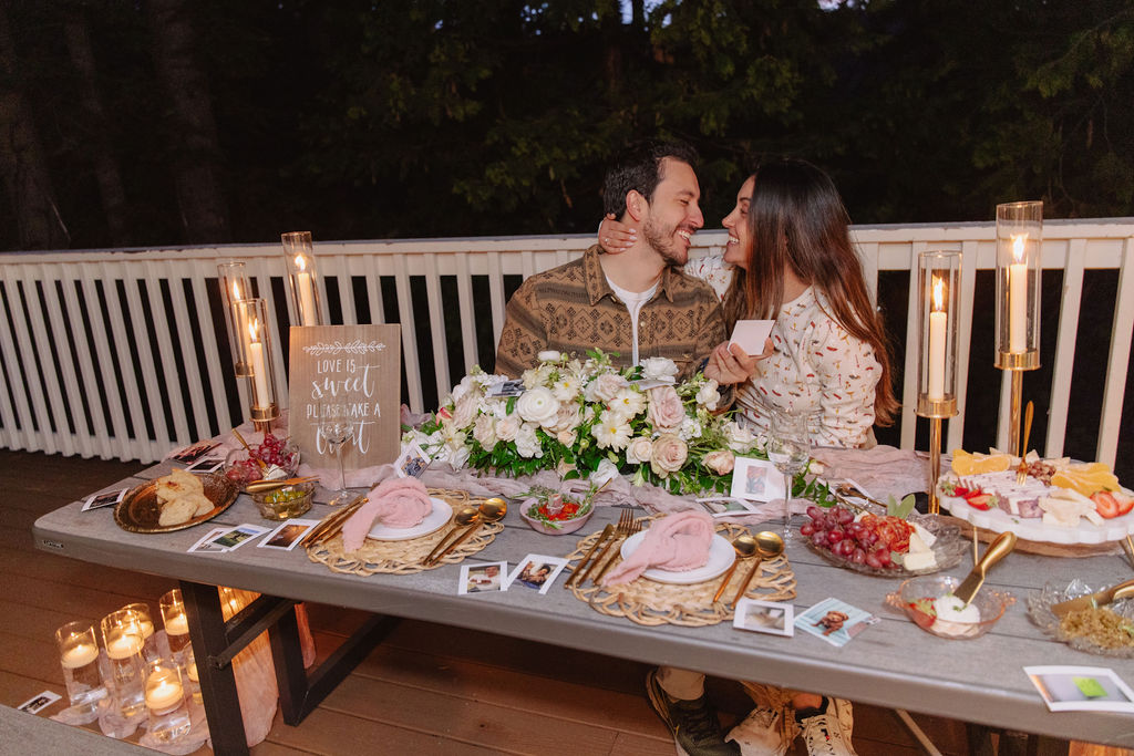 A couple sits on a picnic table, surrounded by candles and flowers, looking at each other. A sign and snacks are in the background.