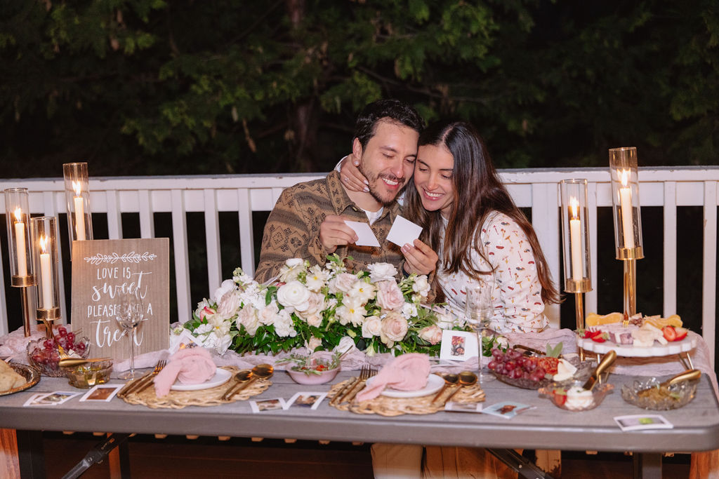 A couple sits at a decorated table with flowers and candles, holding cards and smiling. Plates of food and a board with decorative text are on the table.