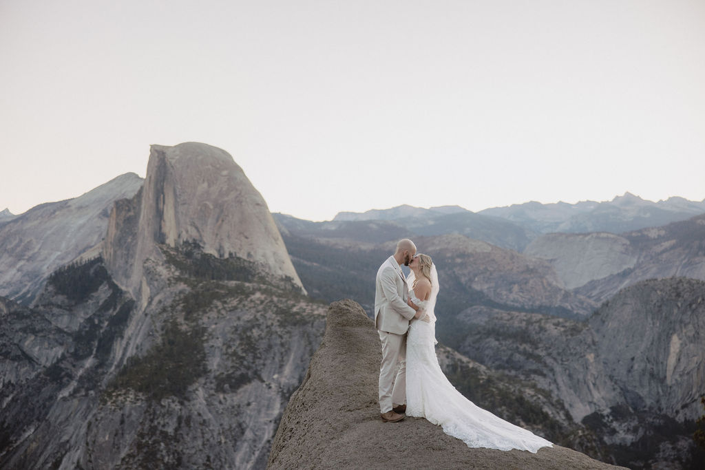 A couple in wedding attire stands on a rocky ledge with a vast mountain landscape in the background. | What to Wear for Your Yosemite Elopement