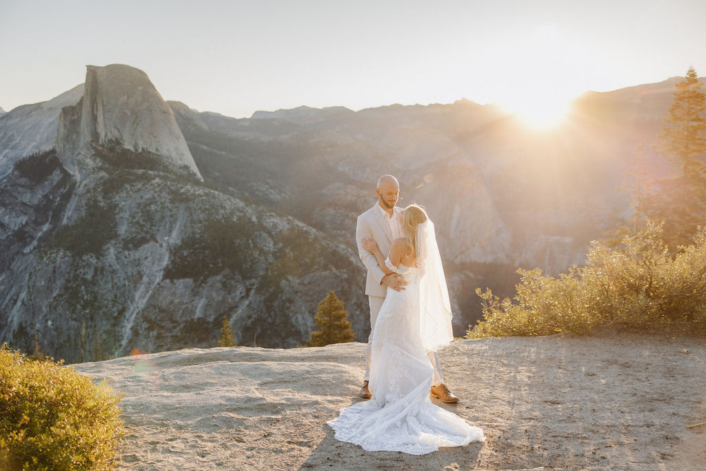 A couple in wedding attire stands on a rocky ledge with a vast mountain landscape in the background. | What to Wear for Your Yosemite Elopement