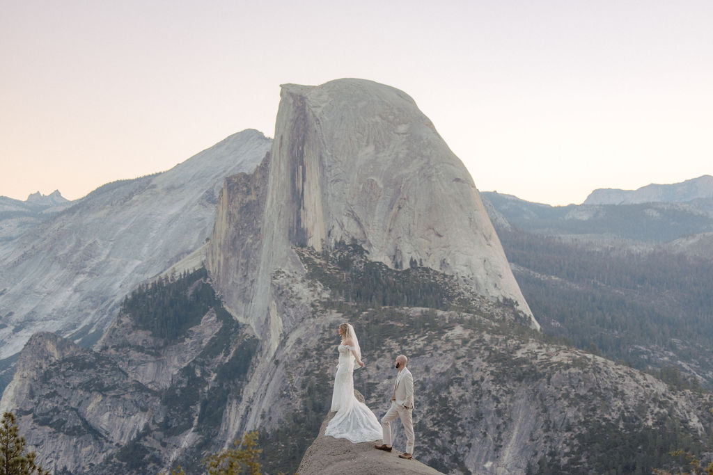 A couple in wedding attire stands on a rocky ledge with a vast mountain landscape in the background. | What to Wear for Your Yosemite Elopement