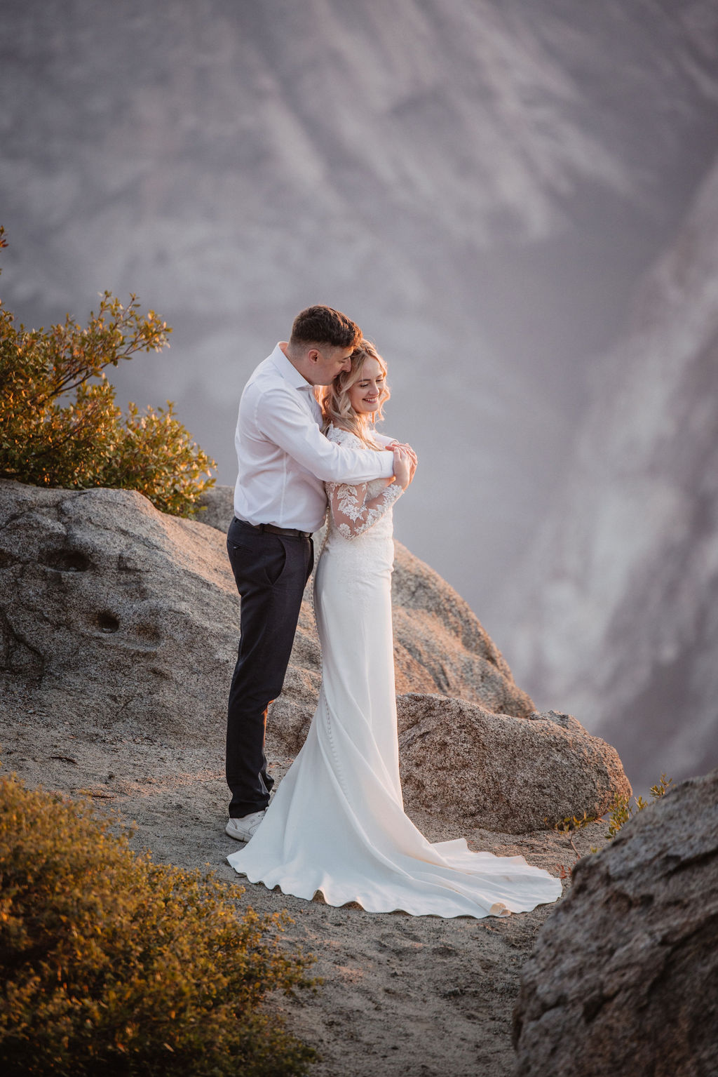 A couple stands on a rocky edge overlooking a vast mountain landscape at sunset.