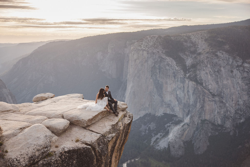 A bride and groom embrace on a rocky cliff with a mountainous landscape in the background under a partly cloudy sky.