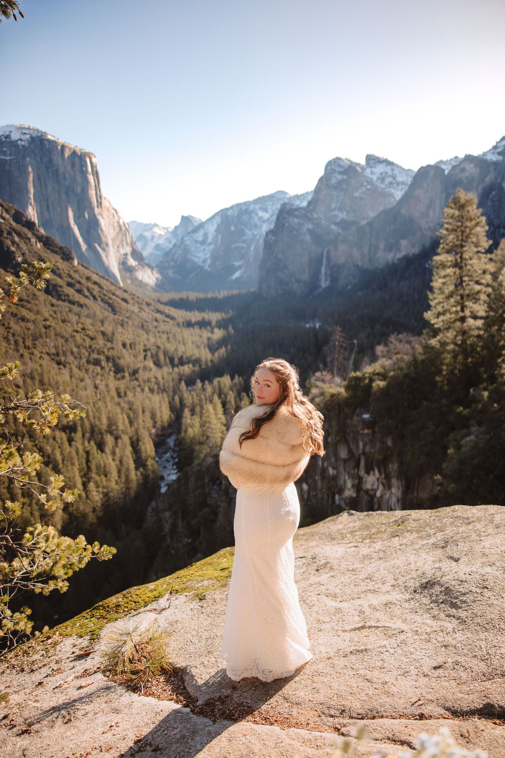 Bride in a white gown and veil stands on a rocky outcrop, with a mountain landscape in the background. |What to Wear for Your Yosemite Elopement