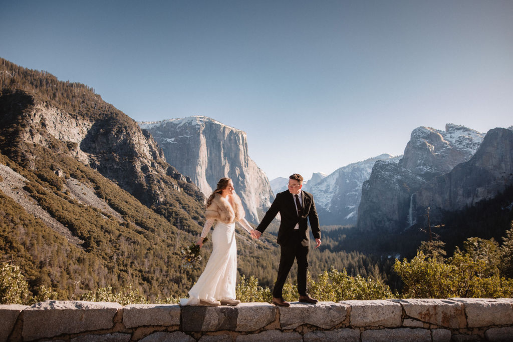 Bride and groom holding hands, standing on a stone wall with a mountainous landscape in the background under a clear sky.