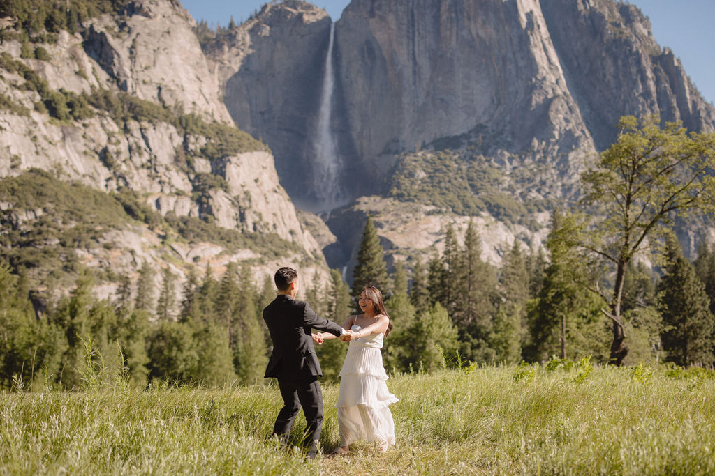 A couple in formal attire dances in a grassy field with a waterfall and mountains in the background.