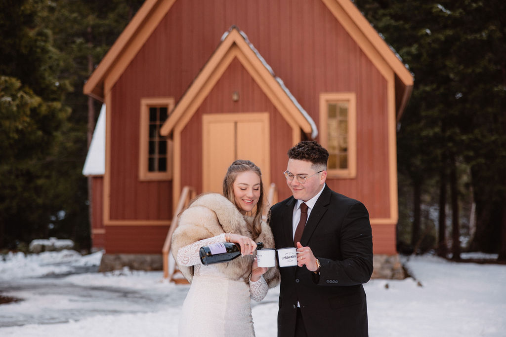 A couple stands in front of a red house in a snowy setting. The woman holds a bottle and pours into a glass held by the man. Both are smiling and dressed in formal wear.