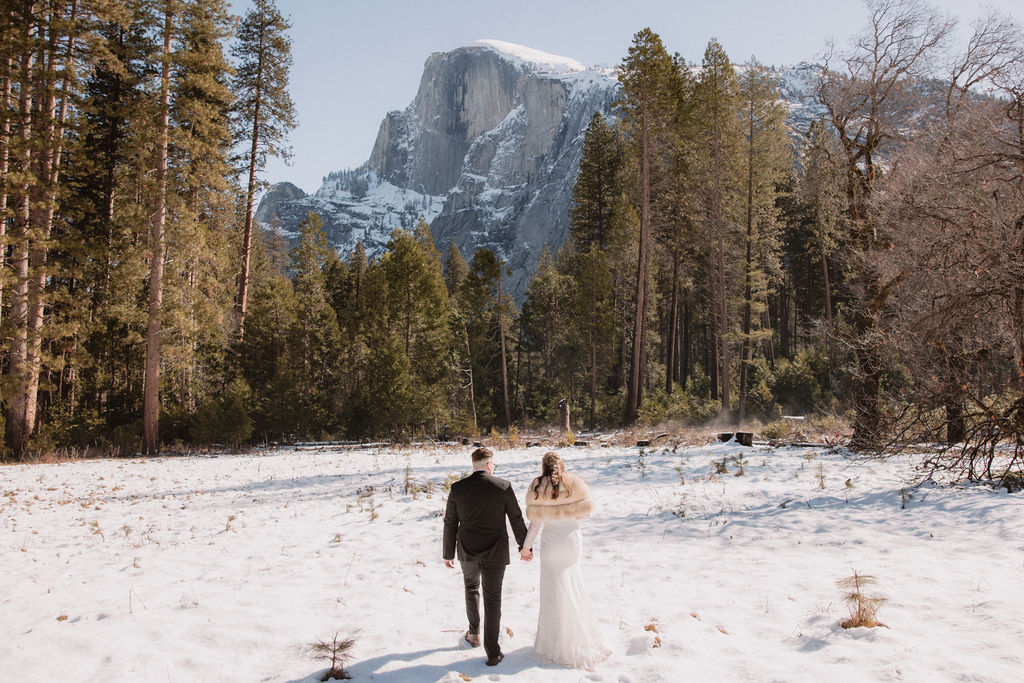 Couple in formal attire holding hands, walking on snow with mountains in the background.