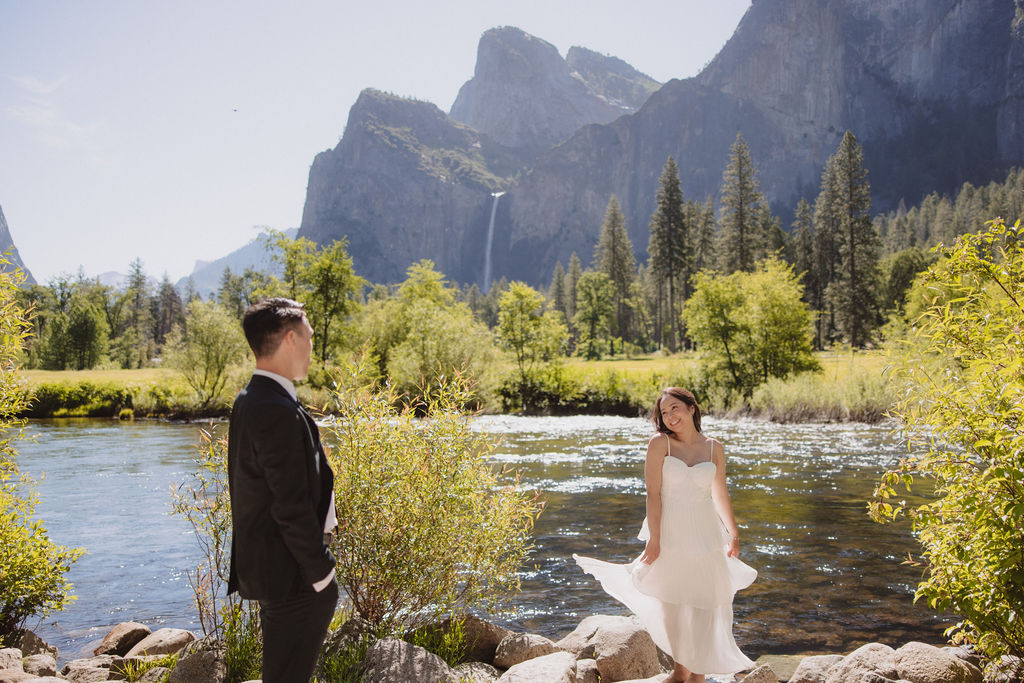 A couple in formal attire dances in a grassy field with a waterfall and mountains in the background.