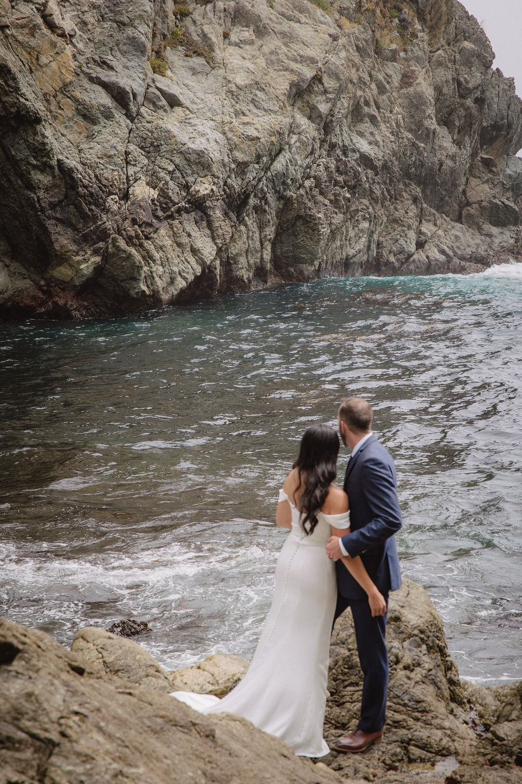 A couple in formal attire poses on rocky seaside terrain with ocean waves and a large cliff in the background for an elopement in California 