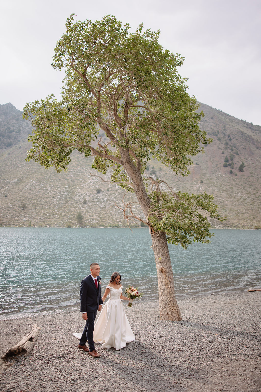 A couple in formal attire walks hand in hand on a rocky lakeside, surrounded by trees and mountains in the background.