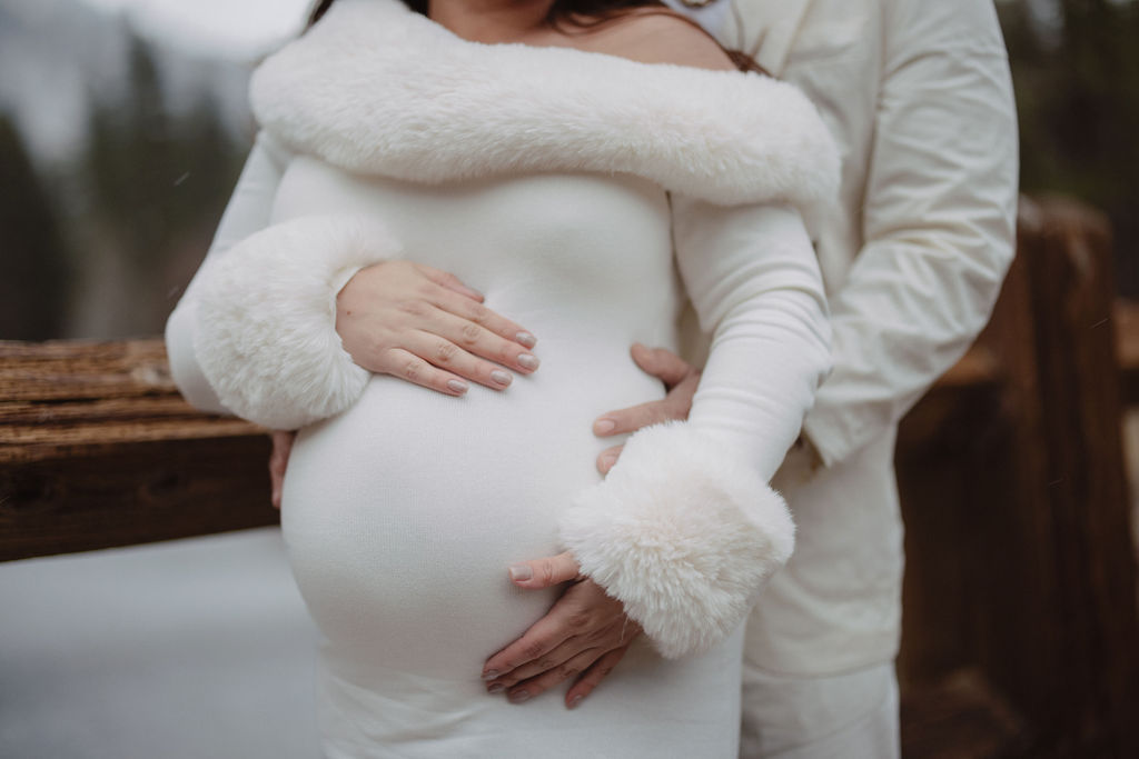 A pregnant person in a white dress stands outdoors, their partner's arms around them, both hands resting on the baby bump.