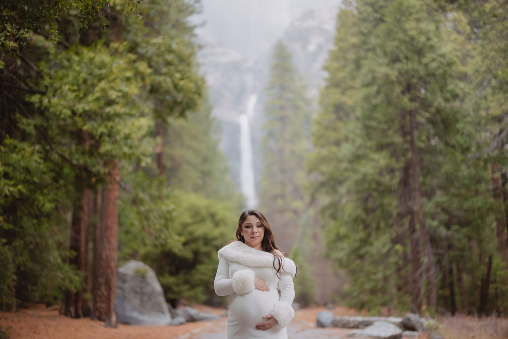 A person with long wavy hair in a white sweater faces away from the camera, reaching up to touch leaves on a tree in a forest setting.