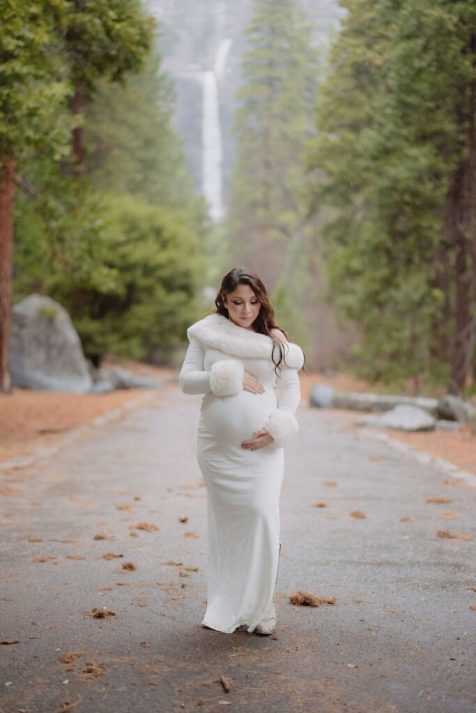 A person with long wavy hair in a white sweater faces away from the camera, reaching up to touch leaves on a tree in a forest setting.