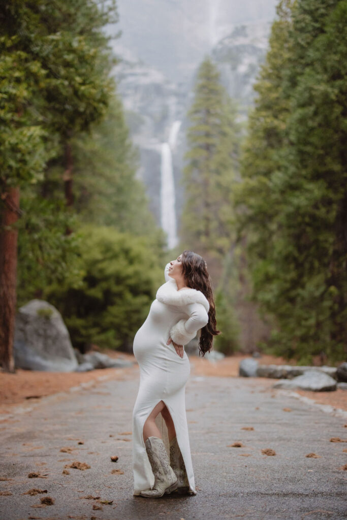 A person with long wavy hair in a white sweater faces away from the camera, reaching up to touch leaves on a tree in a forest setting.