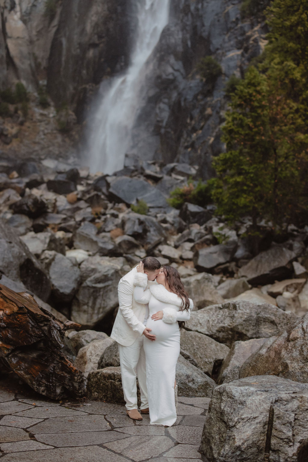 Woman in a white dress stands in front of a rocky landscape with a waterfall in the background.