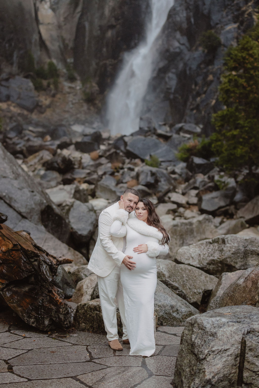 Woman in a white dress stands in front of a rocky landscape with a waterfall in the background.