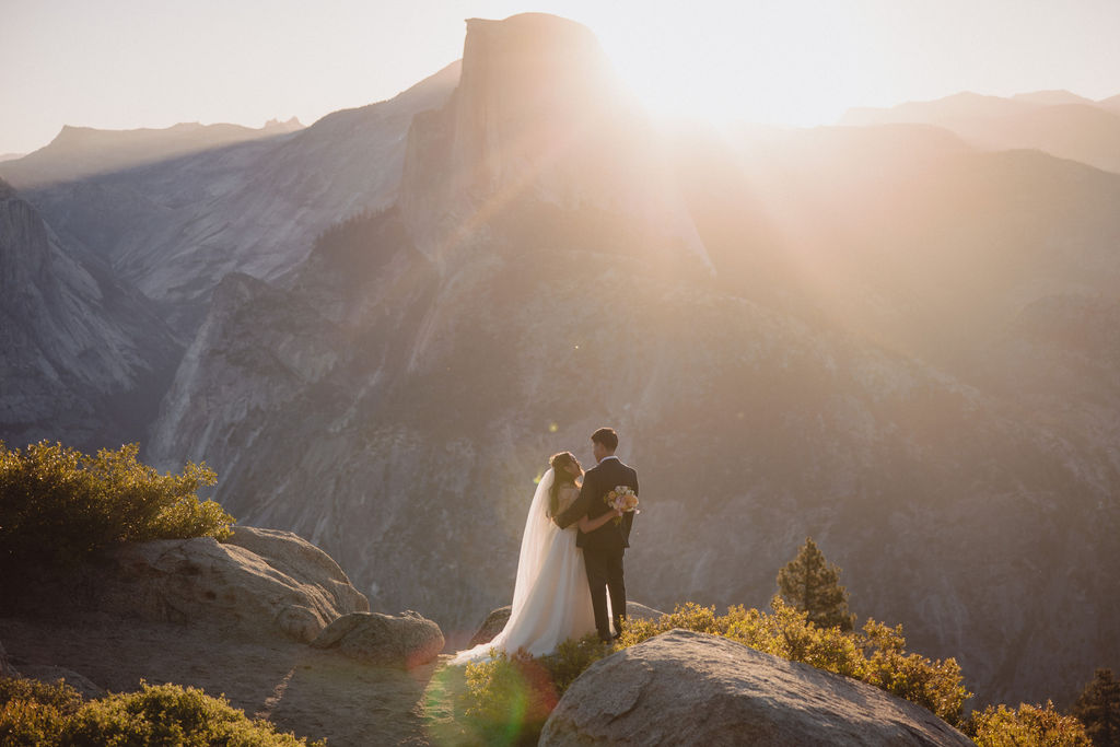 Bride and groom stand on a rocky ledge overlooking a mountainous landscape at sunset.