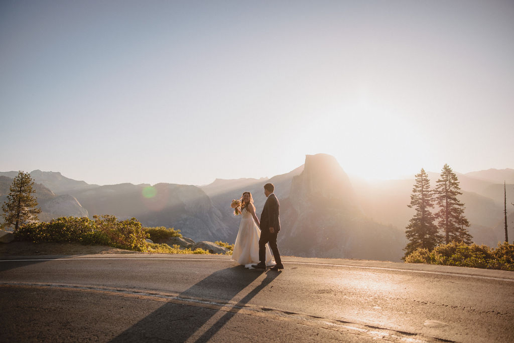 Bride and groom stand on a rocky ledge overlooking a mountainous landscape at sunset.