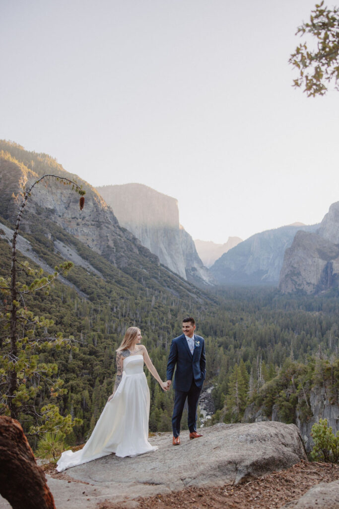  Bride and groom embrace on a rocky ledge overlooking a vast forested valley with towering cliffs in the background. How to Include Friends and Family in Your Elopement