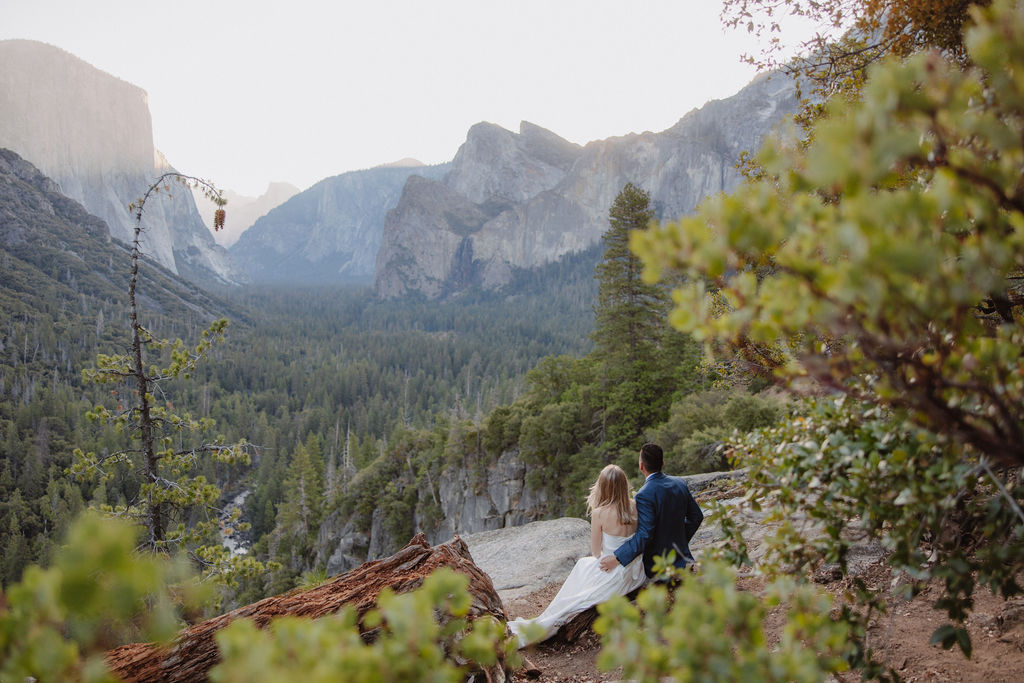 Bride and groom embrace on a rocky ledge overlooking a vast forested valley with towering cliffs in the background. How to Include Friends and Family in Your Elopement