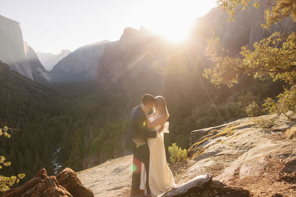 Bride and groom embrace on a rocky ledge overlooking a vast forested valley with towering cliffs in the background. How to Include Friends and Family in Your Elopement