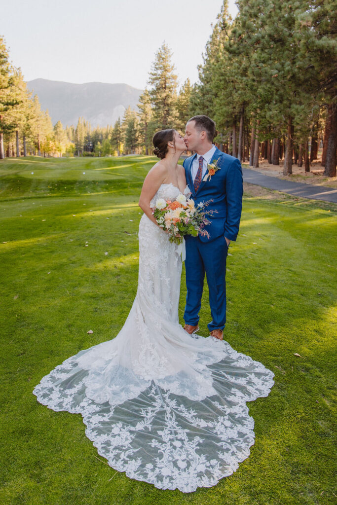 Bride and groom kissing on a grassy field surrounded by trees; bride in a lace wedding dress and groom in a blue suit holding a bouquet for an elopement in california