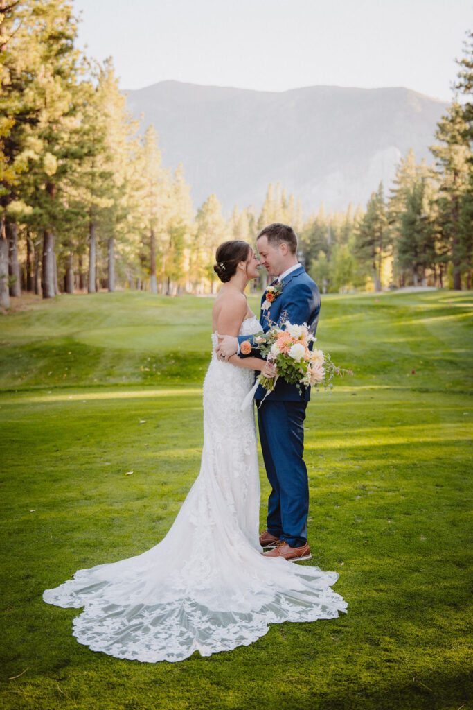 Bride and groom kissing on a grassy field surrounded by trees; bride in a lace wedding dress and groom in a blue suit holding a bouquet for an elopement in california