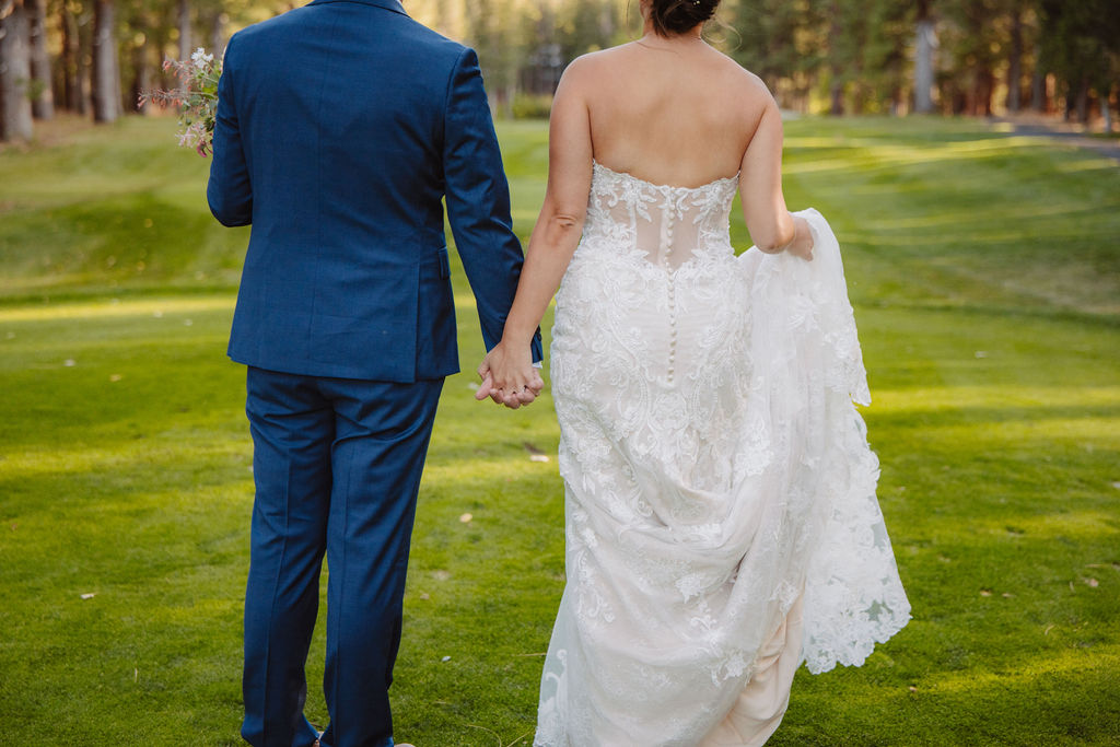 Bride and groom kissing on a grassy field surrounded by trees; bride in a lace wedding dress and groom in a blue suit holding a bouquet for an elopement in california