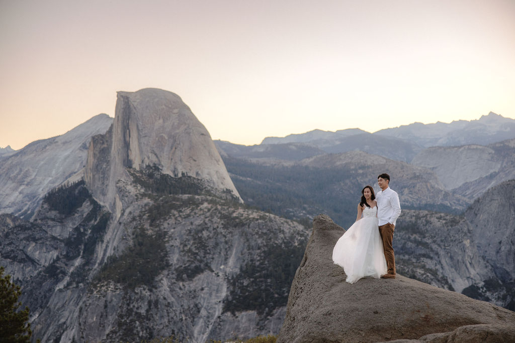 A couple stands on a rocky cliff with a mountain landscape in the background at sunset for their yosemite engagement photos