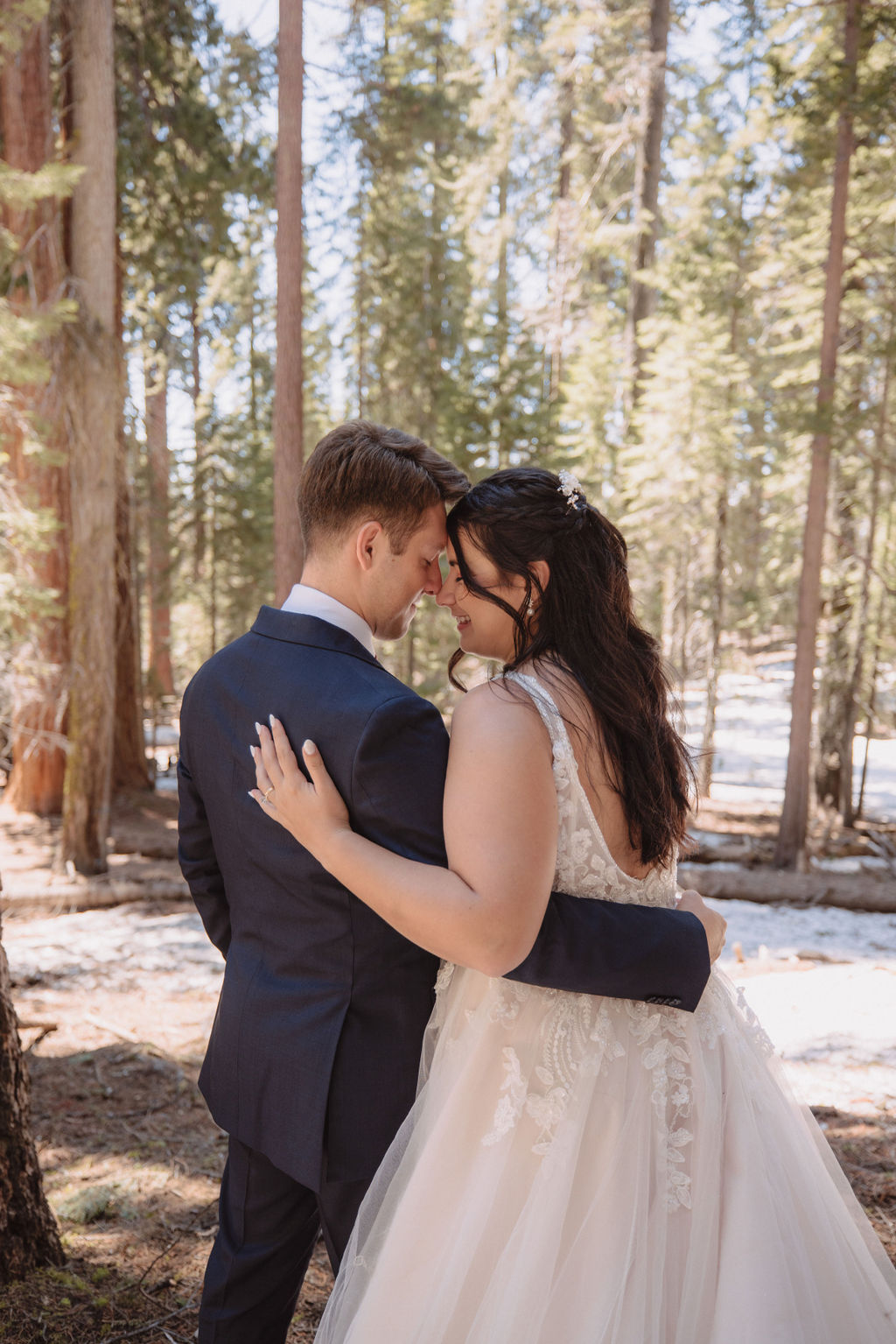 A bride and groom walk hand in hand between two large trees in a forest, with sunlight filtering through the leaves. The bride wears a long white gown. How to Include Friends & Family in Your Elopement